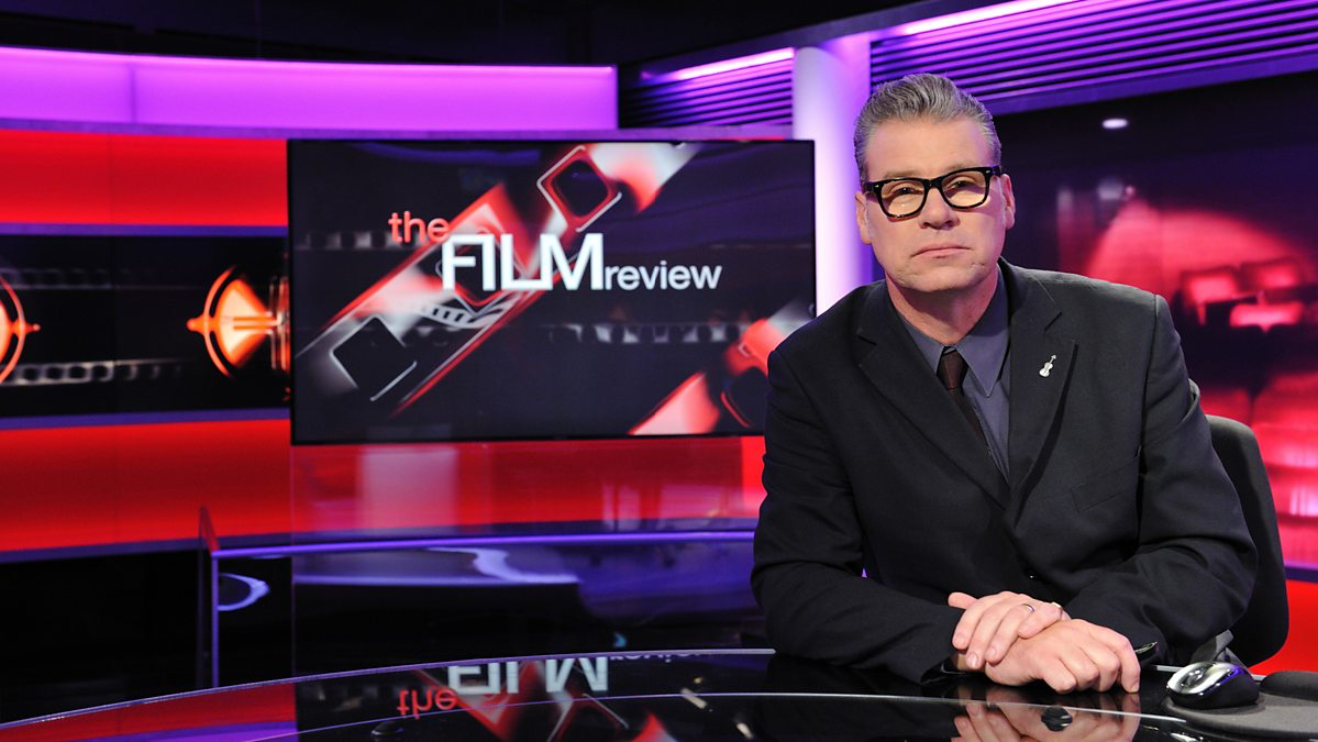 Mark Kermode sat behind a desk looking straight at the camera. A screen is to his right that reads 'The Film Review'.